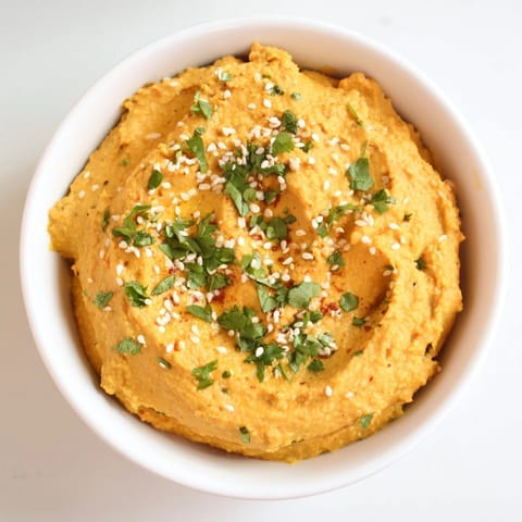 A bowl of creamy Carrot Ginger Dip garnished with sesame seeds and cilantro, served with fresh veggie sticks.