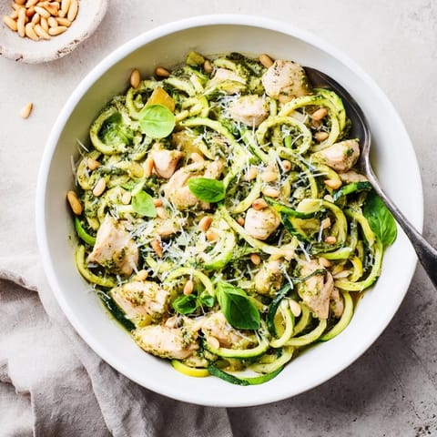 A close-up of pesto zucchini chicken bowl garnished with pine nuts, Parmesan, and fresh basil leaves.